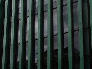 High-contrast architectural detail of a modern building facade, featuring sharp lines and deep shadows, reflecting a dark, overcast sky. Deep forest green and charcoal tones, 35mm.