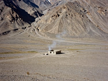 High-altitude landscape of a remote settlement in the Wakhan Corridor, harsh daylight, a single stone dwelling with smoke rising, minimalist composition.