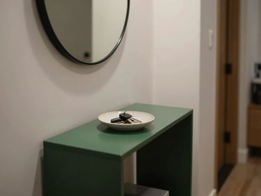 Close-up of a narrow apartment entryway, a slim dark green console table holding a ceramic dish for keys, a round mirror reflecting soft light, minimalist styling.