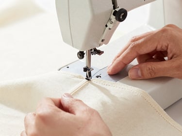 Macro detail of a high-end industrial sewing machine stitching a clean seam on ivory cotton canvas, hands of the artisan visible, bright natural studio lighting.