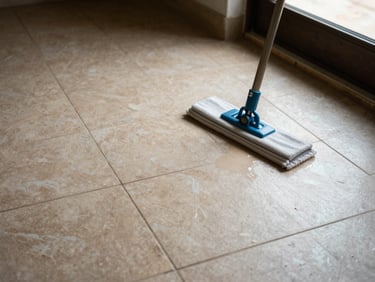 Overhead detail of a freshly cleaned tiled floor, mop resting to the side, morning light entering from a window, warm surface tones, documentary style
