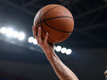 Low-angle action shot of a player mid-drive under dramatic stadium lighting, hand gripping a worn basketball, high-contrast shadows.