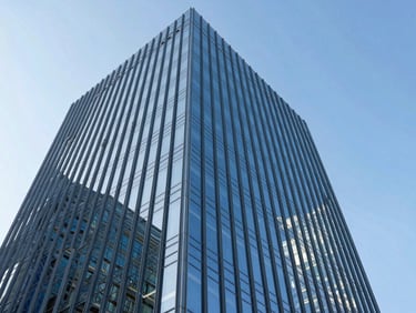 Low-angle architectural photography of a towering modern glass facade reflecting a bright blue sky, sharp daylight, steel blue tones, professional tilt-shift lens