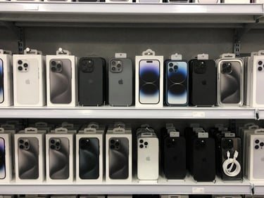 Overhead of assorted mobile accessories — phone cases, screen protectors, charging cables in retail packaging — arranged in neat rows on a light grey warehouse shelf, even lighting, no people