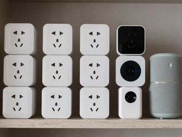 Overhead close-up of smart home devices — smart plugs, mini speakers, tracking devices — arranged in organized grid on a neutral shelf under studio lighting, no people
