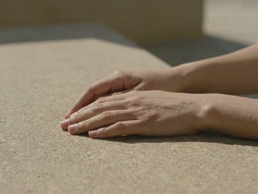 Close-up of gentle hands resting on a warm-toned textured stone surface, soft architectural shadows, muted olive and stone tones, shallow depth of field, 35mm photography
