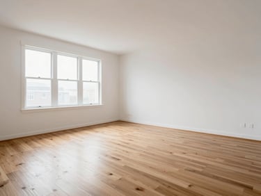 Wide shot of an empty residential unit interior, bare hardwood floors under flat window light, clean white walls, no furniture or staging, architectural condition clearly visible