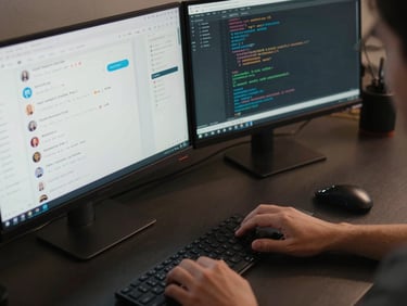 Close-up overhead view of a developer's desk — dual monitors displaying a chatbot admin panel and a code editor with API route definitions, keyboard hands mid-type, cool studio lighting, dark desk surface