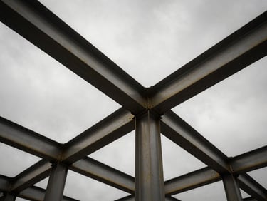 Low-angle heroic perspective of structural steel beams rising into a dramatic overcast sky, high-contrast desaturated industrial photography.