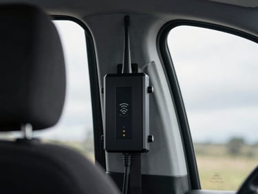Interior of a van showing a cellular signal booster unit mounted on the wall, a single antenna cable running upward, overcast window light, close-up framing showing the device label and indicator LEDs