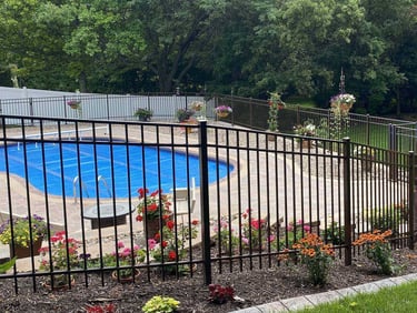 In-ground swimming pool with a blue solar cover enclosed by a black metal security fence and floral landscaping.