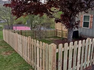 A wooden picket fence encloses a green backyard lawn with a red maple tree near a residential house.