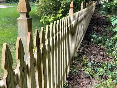 Wooden picket fence with gothic tops installed along a lush green backyard garden.