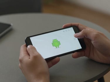 Tight crop of fingers holding an Android phone showing a simple game screen, resting on a coffee table under neutral indoor light