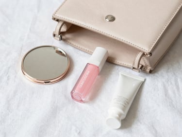 Aesthetic flatlay of an open beige leather handbag showing a pastel pink lip oil, a compact mirror, and a small hand cream on white linen