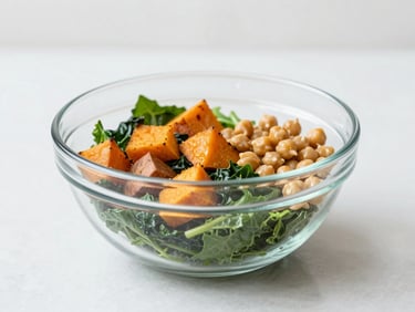Bright minimalist photo of a large glass bowl containing dark leafy greens, roasted sweet potato cubes, and chickpeas under diffused natural light.