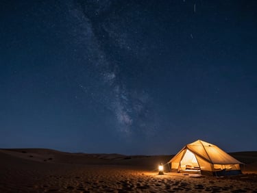 A wide low-angle shot of a luxury desert camp under a brilliant night sky thick with stars, warm lantern light glowing from within a single tent in the right third, deep indigo sky dominating the frame.