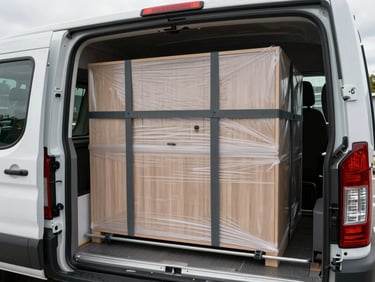 Interior of a medium panel van loaded with wrapped furniture, straps securing a wardrobe against the side wall, overcast daylight from rear opening, visible tie-down rails along the floor
