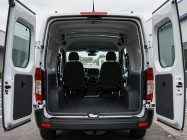Interior of a compact cargo van showing loading space, overcast daylight through rear doors, empty clean cargo area visible, vehicle parked at a building entrance