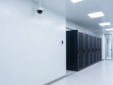 Wide angle environmental shot of a modern hospital corridor with clean white walls, featuring an integrated dome CCTV camera and access control panel mounted near a glass door, cool lighting.