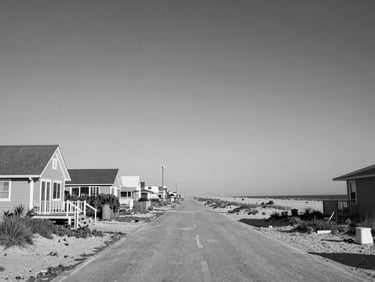Black and white wide shot of the Surfside Beach waterfront, modest beach cottages along a quiet street, ocean visible in background, natural daylight, low horizon