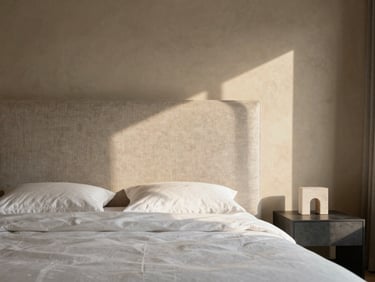 Wide shot of a high-end bedroom interior, linen upholstered headboard against a warm plaster wall, side table with a single architectural object, naturalistic morning light from the right, no people