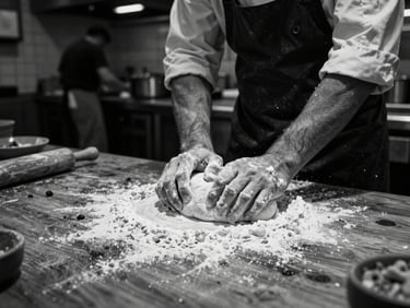 Wide-angle, high contrast black and white photo, low-key ambient light, showing a chef's hands covered in flour, powerfully kneading dough on a rough wooden table in a busy, industrial kitchen.