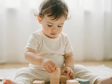 Candid mid-shot of a two-year-old toddler playing with wooden blocks, wearing a soft cream cotton set, diffused morning daylight.