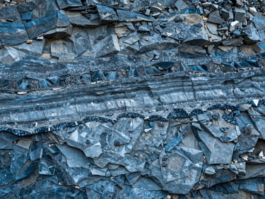 Overhead wide study of exposed quarry strata wall, geological layers in cool blues and greys stratified horizontally, raw stone surface showing mineral veins and crystalline formations, overcast diffused light, no human presence, no color correction