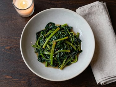 Close-up overhead flatlay of a restaurant place setting — a shallow ceramic bowl with braised greens, a linen napkin folded beside it, candlelight edge glow on a dark wood surface, north-facing daylight from the left
