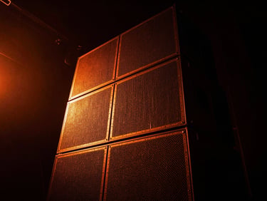 Dramatic low-key photo of a massive speaker stack on a dark stage, vibrant electric orange light leaks, gritty texture, wide angle.