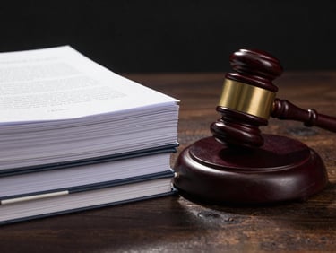 Close-up of a judge's gavel resting beside a stack of bound legal briefs on a dark wood surface, tungsten desk lamp illuminating the document spines, deep shadow behind, no people, precise and still