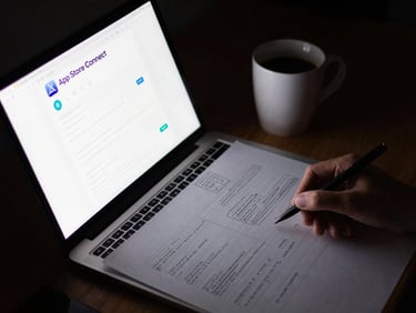 Overhead shot of a desk with a laptop displaying App Store Connect, a hand-drawn UI wireframe on paper, and a coffee cup, moody studio lighting with subtle reflections