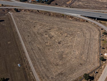 Aerial photograph of a raw land parcel adjacent to a highway interchange, visible on-ramp infrastructure at the parcel edge, flat terrain with clear boundary markers, crisp daylight, wide environmental framing with highway in the upper third