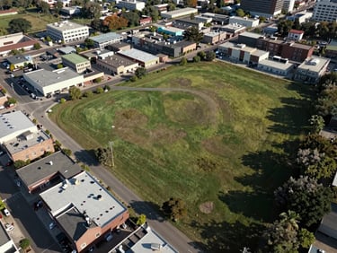 Wide aerial view of a semi-urban infill land parcel bordered by existing low-rise commercial structures, access road visible on the western edge, natural daylight casting long shadows, broad environmental framing with the parcel occupying the right half