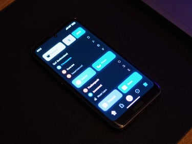 Moody tech photography, low-key ambient lighting, vibrant neon screen glows, sharp macro details, 35mm lens. An overhead flat-lay of an Android device showing a sleek dark-mode dashboard UI with sharp neon-blue accents, resting on a clean dark desk.