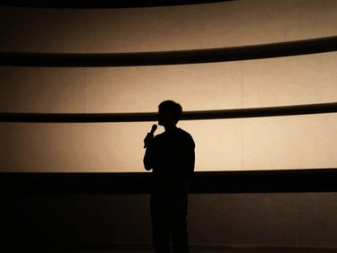 Low-key dramatic portrait of a speaker silhouetted against a warm golden-hour stage light, architectural lines of a modern amphitheater, cinematic atmosphere