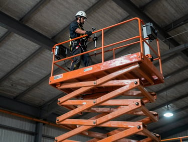 An orange scissor lift extended high near an industrial ceiling, operator wearing full fall-protection harness and hard hat.