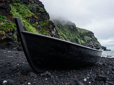 A close-up, high-contrast shot of a heavy black zodiac boat resting on the rocky, volcanic shoreline of a remote island, with a steep, green cliffside rising into the mist behind it.