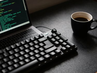 Tight macro shot of a clean dark workspace, a mechanical keyboard, code on a laptop screen reflecting terminal green, and a single-origin espresso in a matte black cup under sharp directional window light.