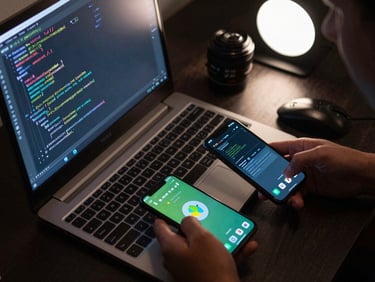 Close overhead shot of a developer's keyboard and two Android phones mid-session on a dark work table, code visible on the nearest screen, studio strobe light rendering the screens clearly without glare, hands partially in frame