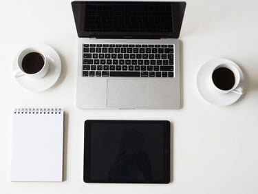 Structured overhead desk shot with modern tech tools (laptop, tablet, notepad) and a coffee, clean minimalist background, natural daylight