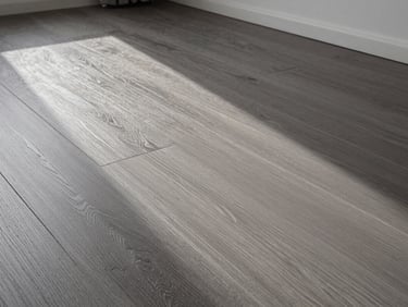 Wide-plank engineered wood floor detail in a luxury apartment, cool north daylight raking across the grain, precise baseboard joint visible, grey tones, no people