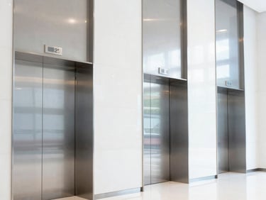 Crisp architectural photography of a modern commercial office lobby, bright daylight reflecting off polished steel elevator doors, cool undertones.