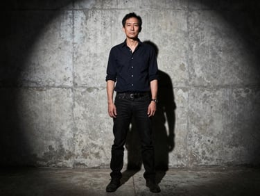 Stark environmental portrait of the creator standing in a concrete warehouse, harsh direct flash, dramatic shadows.