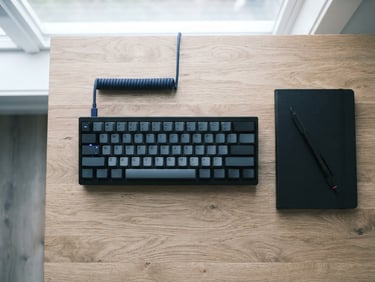 Overhead flat-lay of a minimalist wooden desk setup, a clean mechanical keyboard, a black notebook, and a single mechanical pencil under soft natural light, desaturated tones with a subtle blue accent, 35mm