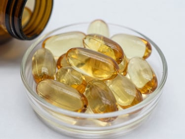 Close-up of omega-3 fish oil capsules in a clear glass dish on a white surface, cool studio lighting from above, capsules in sharp focus showing amber translucency