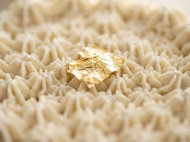 Macro shot of a pristine, hand-piped vanilla bean cake texture with a single edible gold leaf, soft morning light, shallow depth of field