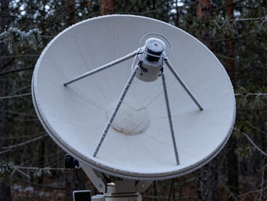 Extreme close-up of a ruggedized satellite dish covered in light frost, mounted on a solid steel bracket against a dark pine forest background.