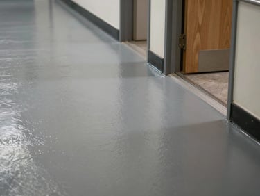 Close-up overhead shot of epoxy floor in a school hallway, mid-grey surface under fluorescent light, showing wear-resistant texture near a doorway threshold, no people, institutional ambient light